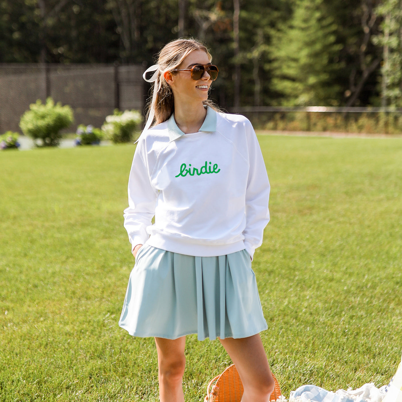 Woman stands on grass wearing a white sweatshirt that has the word birdie embroidered on the front of it in green