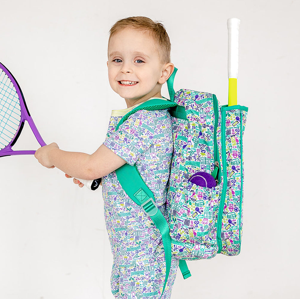 Little boy wearing shirt and pants pajama set. Pajamas have hand drawn tennis pattern with tennis balls, net and racquet in green yellow and purple along with matching kids tennis backpack