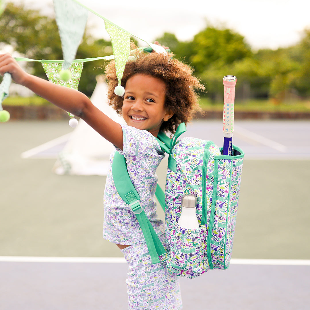 little girl wearing shirt and pants pajama set. Pajamas have hand drawn tennis pattern with tennis balls, net and racquet in green yellow and purple. She is also wearing the match tennis backpack
