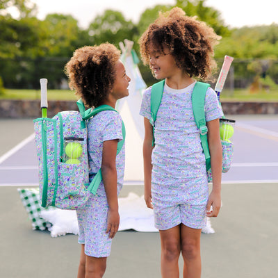 sisters stand on tennis court wearing shirt and pants pajama set. Pajamas have hand drawn tennis pattern with tennis balls, net and racquet in green yellow and purple.