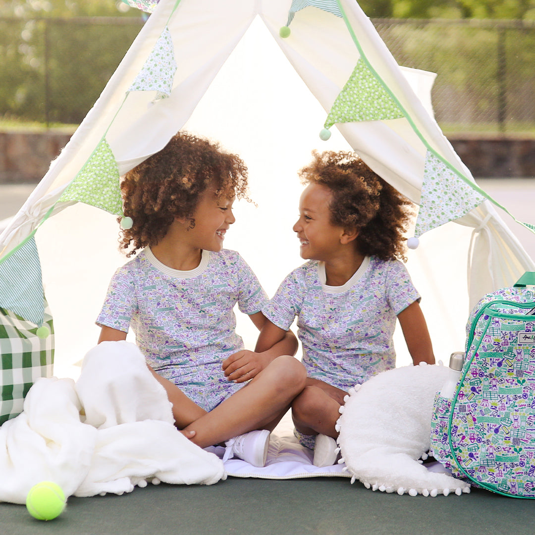 sister sit under white tent wearing shirt and pants pajama set. Pajamas have hand drawn tennis pattern with tennis balls, net and racquet in green yellow and purple.