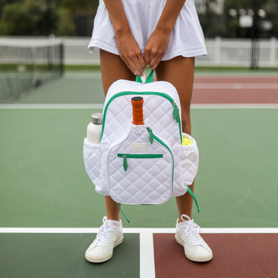 Model stands on pickleball court holding pickleball backpack with white quilted nylon fabric. Backpack has green trim and front paddle pocket.