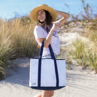 Model stands on beach holding white nylon beach tote with navy straps and navy details