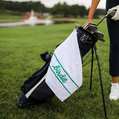 White cotton terry towel with the word "birdie" embroidered in a green cursive font on the bottom of the towel clipped to a golf bag on a carabiner 