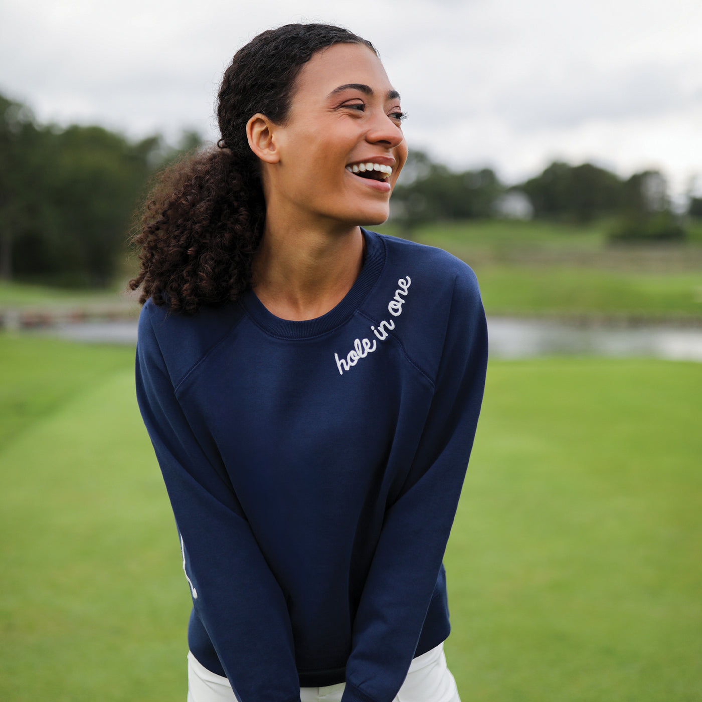 Navy womens cotton sweatshirt with "hold in one" in white cursive font embroidered on the neckline and white crossed golf clubs embroidered on the elbows.