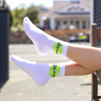 woman sits on a tennis court with her legs in the air. She wear a pair of white socks with a lime green tennis ball and the word love stitched around the ankle