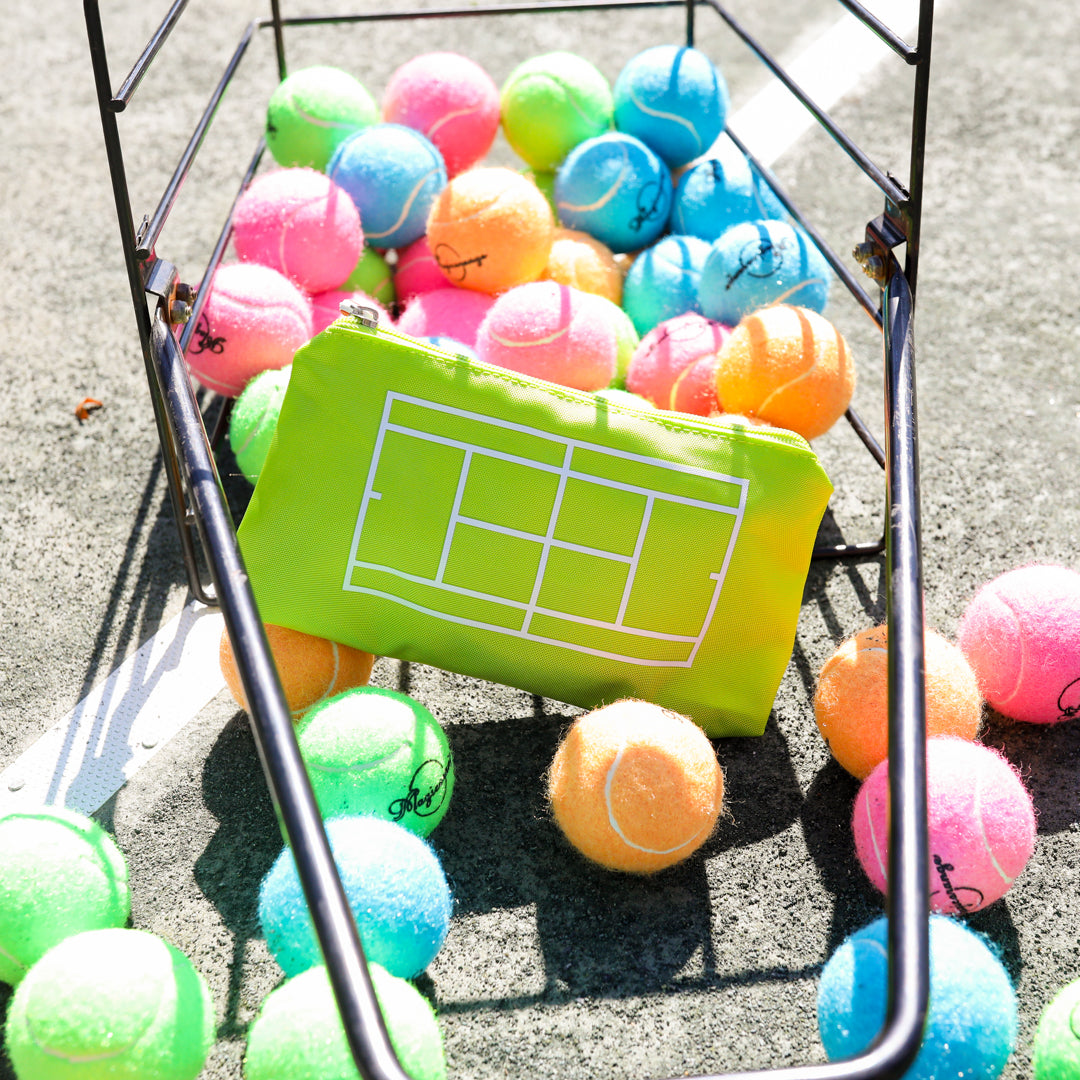 Basket of tennis balls is tipped over on a tennis court with florescent yellow everyday pouch inside basket. Pouch has white tennis court printed on front and red tennis racquet zipper pull.