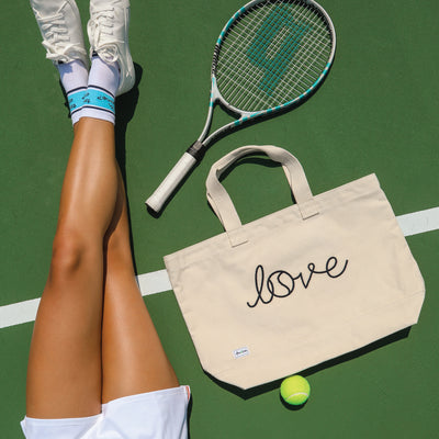 Woman lays on tennis court with Canvas large tote with the front embroidered with the word love in a navy cursive font