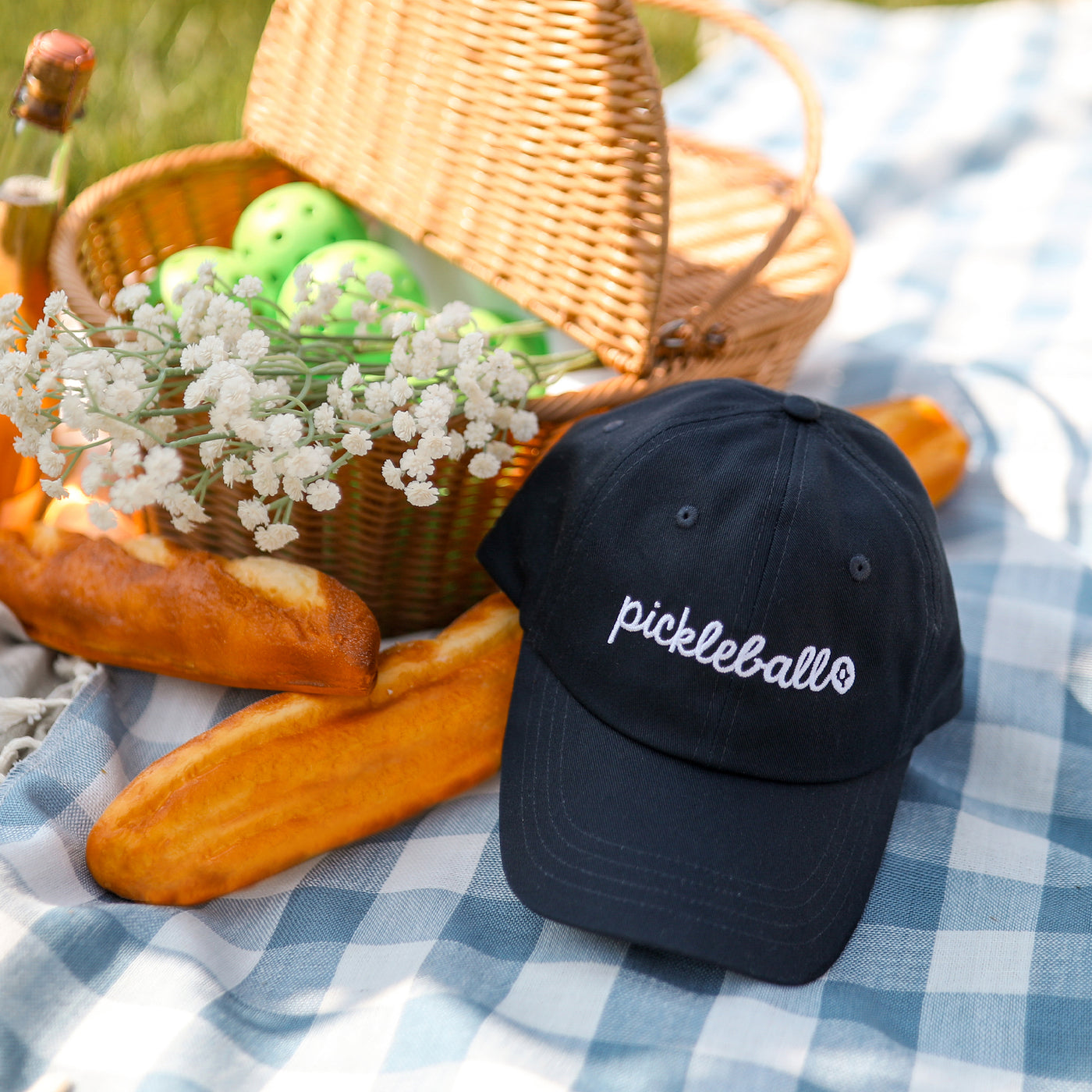 Navy baseball hat on picnic blanket next to basket full of pickleballs. Hat is embroidered with the word pickleball