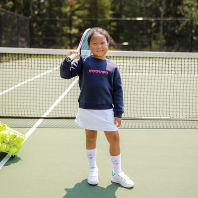 Little girl stands on a tennis court with a tennis racket over her shoulder. She is wearing a navy sweatshirt with pink hearts embroidered on the front