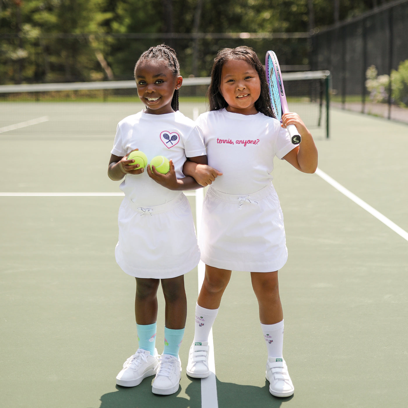 Two little girls stand on a tennis court. Both are wearing white t shirts. One is embroidered with a pink heart and navy racquets. The other is embroidered with the words tennis anyone in pink cursive front