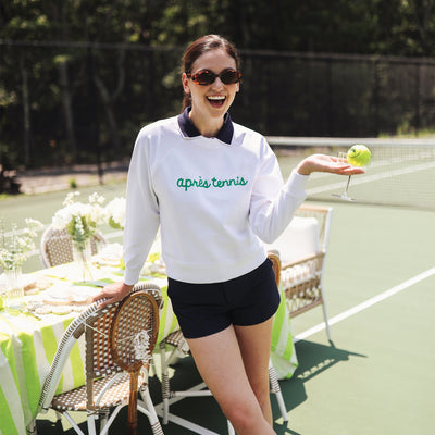 Woman stands on a tennis court laughing wearing a white sweatshirt with the words "apres tennis" embroidered on the front in green cursive