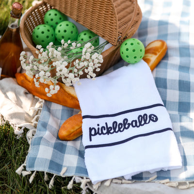 White terry towel laying flat on white background with navy stripes and the word pickleball embroidered on it. Towel is on a picnic blanket next to a basket