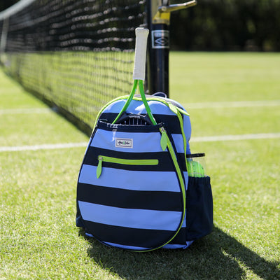 front view of navy and blue striped kids tennis backpack with green trim and zippers. Tennis backpack is on the ground in the grass next to a tennis net