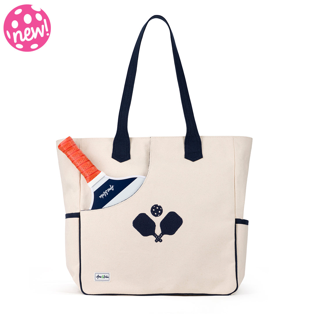 Front view of natural canvas baseline pickleball tote. Tote has navy trim and handles. Embroidered navy crossed paddles on the front. Front slip pocket is holding a pickleball paddle