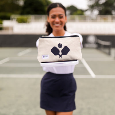 Model holds out paddles with makeup case on it. front view of small natural canvas makeup pouch. Navy crossed paddles are embroidered on the front and navy trim on the zipper.