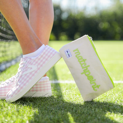 Model stands with Small natural canvas pouch with lime green zipper and embroidered word "matchpoint" in lime green on fron