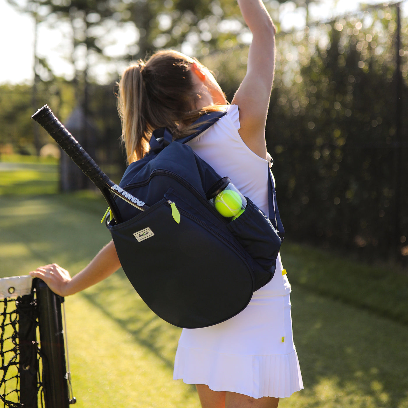 Model stands on grass tennis court wearing navy tennis backpack.