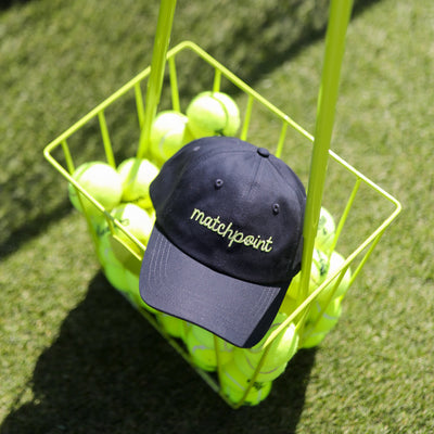 Navy womens baseball hat with lime embroidery on the front that reads "matchpoint" sits in a tennis basket on a tennis court