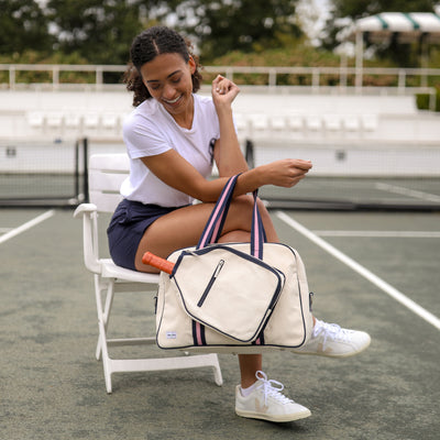 Model sits on pickleball court holding natural canvas pickleball tote with navy trim and navy and pink straps and handles.