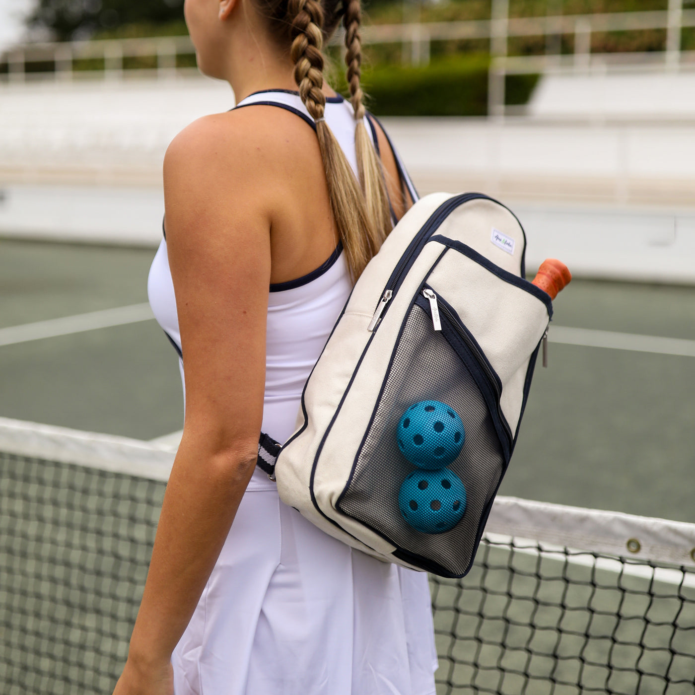 Model wears Hamptons pickleball sling bag. Bag has front mesh pocket and slip pocket on front to hold pickleball paddles. Bag has navy trim and navy and white striped straps. Bag has adjustable straps to be worn as a backpack or a crossbody.