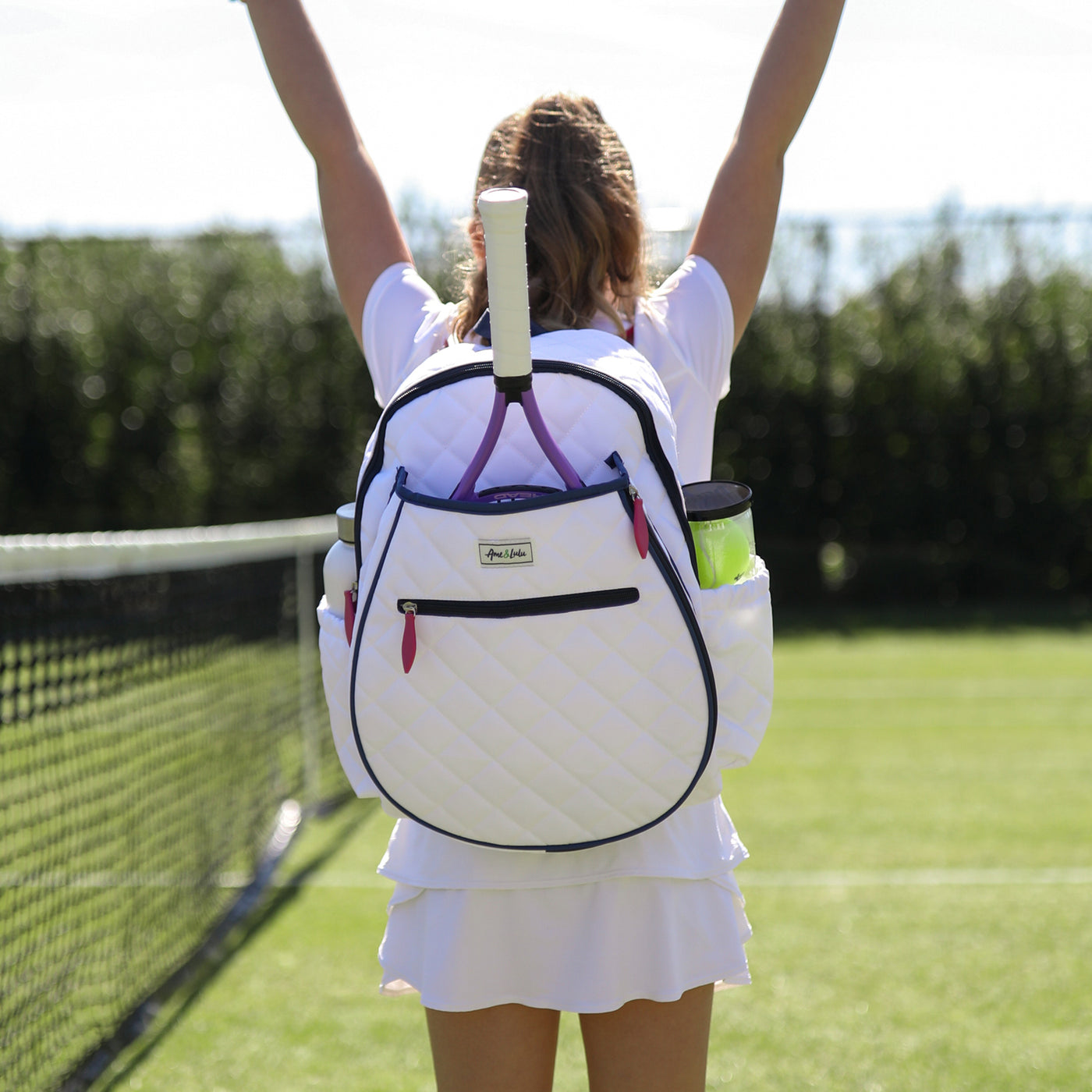 girl stands on tennis court wearing quilted white kids tennis backpack with navy trim and hot pink zippers.