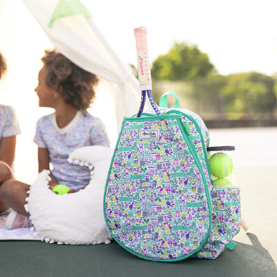 joy street collab kids tennis backpack sits on tennis court next to two little girls. Bag has drawings of tennis balls racquets and nets in green yellow and purple on the bag