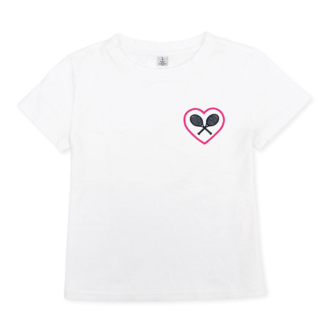 White kids t-shirt lays flat on a white background. There is a pink heart with navy crossed racquets inside embroidered on the pocket area of the shirt