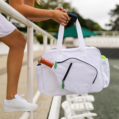 model holds white pickleball tote with navy trim and green zipper pulls. Tote had front paddle pocket.