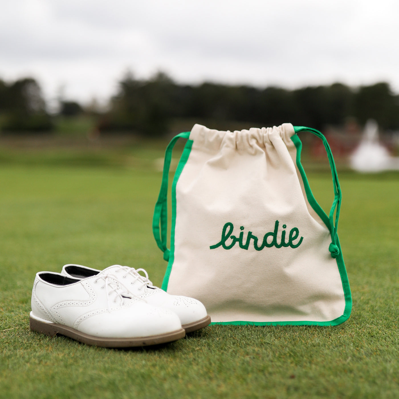 front view of natural canvas shoe bag with green trim and the word "birdie" embroidered on the front in green.