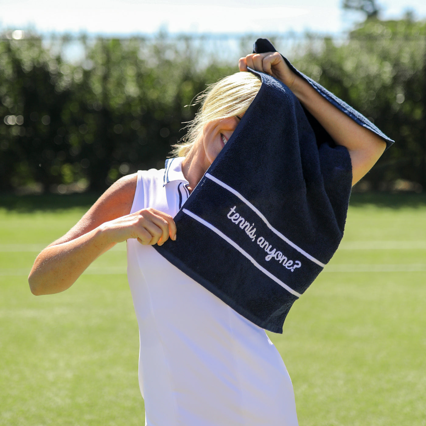 Model stands on tennis court holding Navy cotton terry towel with the words "tennis anyone" embroidered in a cursive font in white on the bottom of the towel.