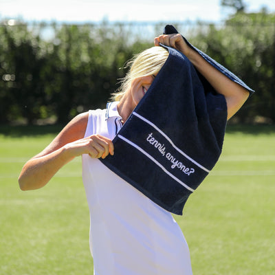 Model stands on tennis court holding Navy cotton terry towel with the words "tennis anyone" embroidered in a cursive font in white on the bottom of the towel.
