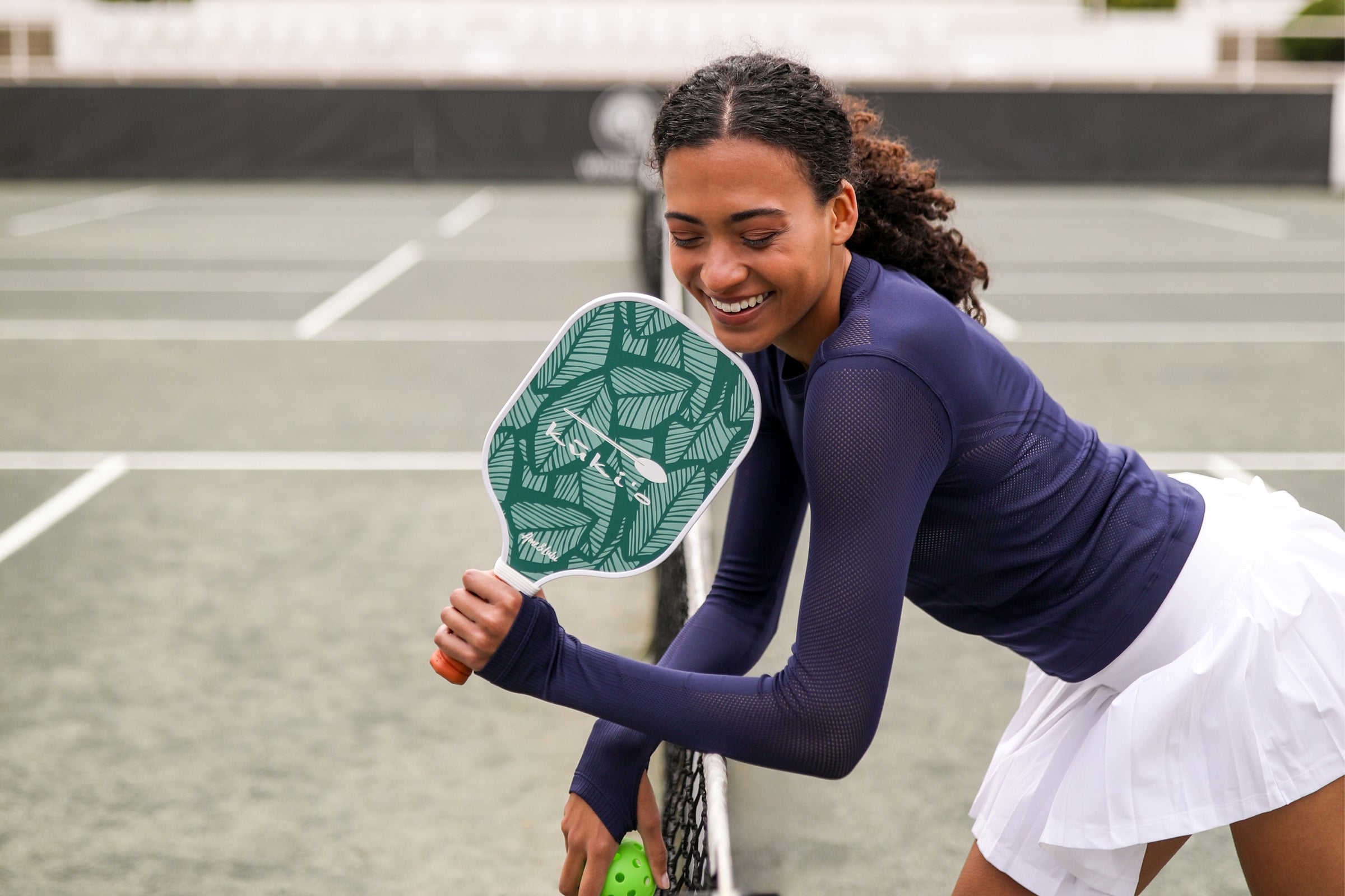 A smiling woman in athletic wear, adorned with active lifestyle accessories, playing pickleball on an outdoor court.
