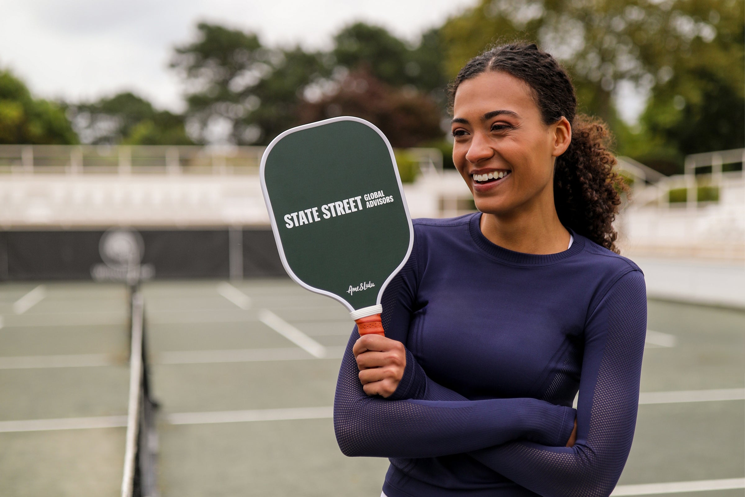 Smiling woman with a state street tennis paddle on a court exudes positivity.