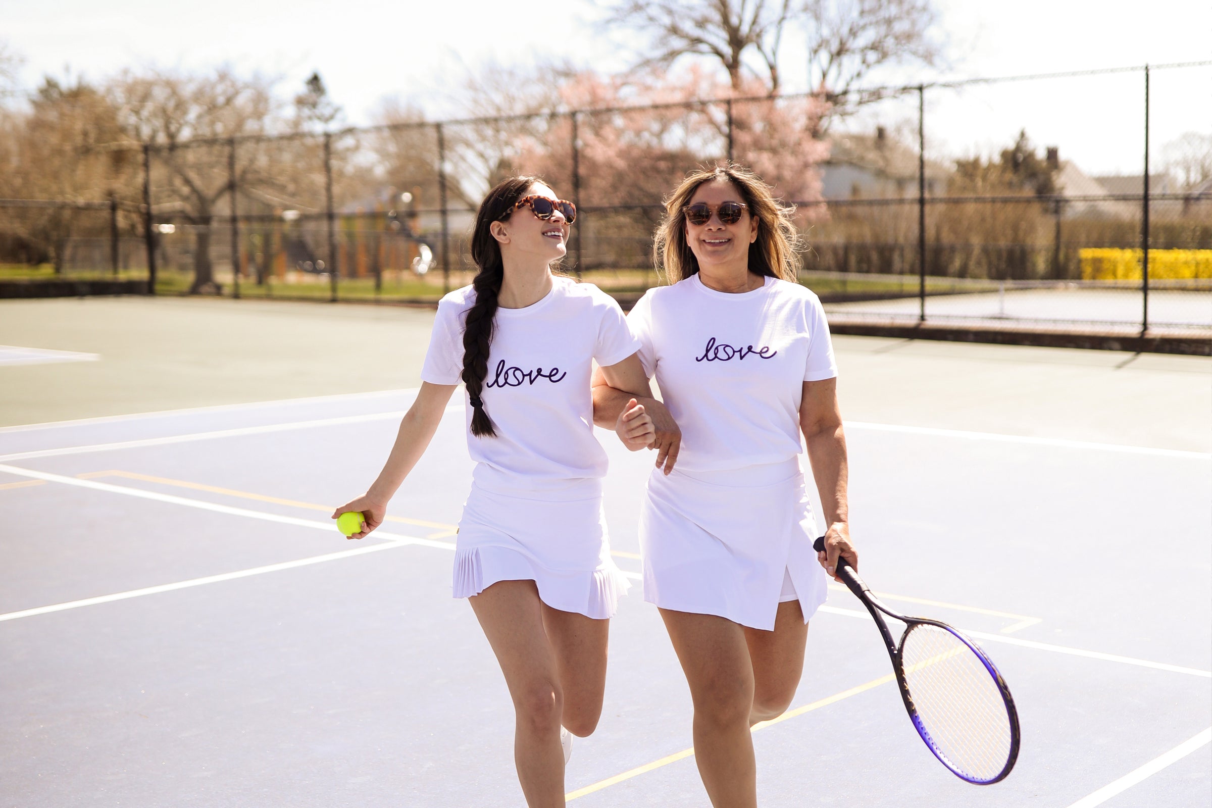 Mother and Daughter walk and laugh together on a tennis court. They are both wearing white t-shirts that say "love" in navy cursive font.