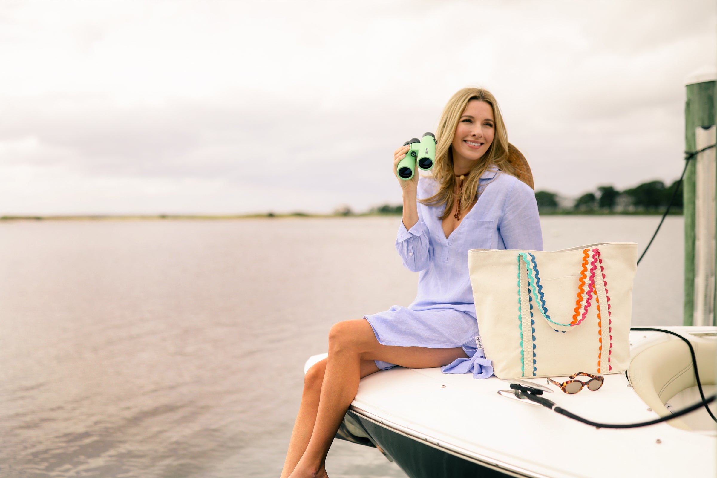 Woman on a boat holding binoculars with a colorful tote bag beside her