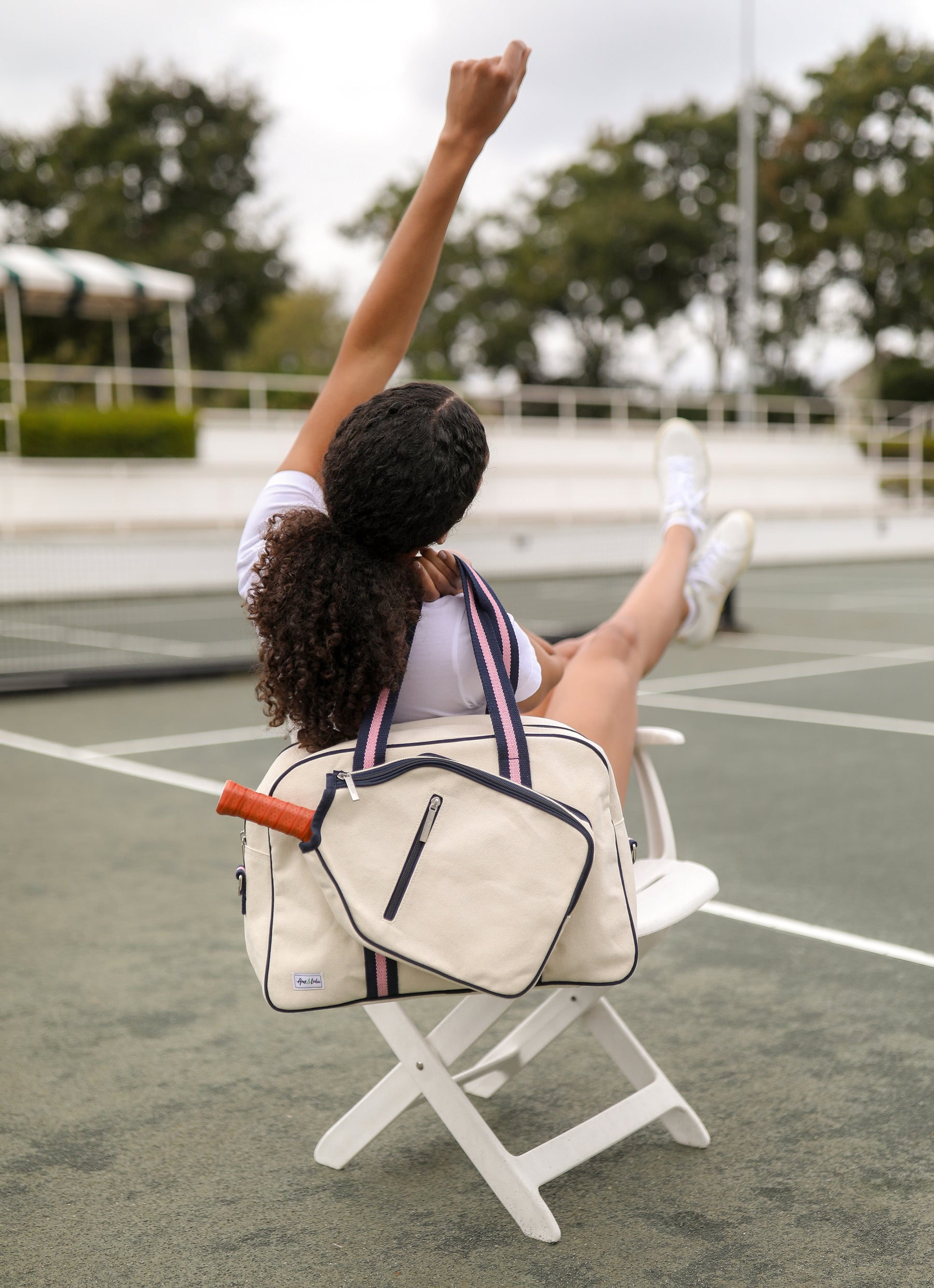 woman sits in a white chair on a pickleball court. She has one arm up in the air and the other is holding a canvas pickleball tote with navy and pink trim over her shoulder.