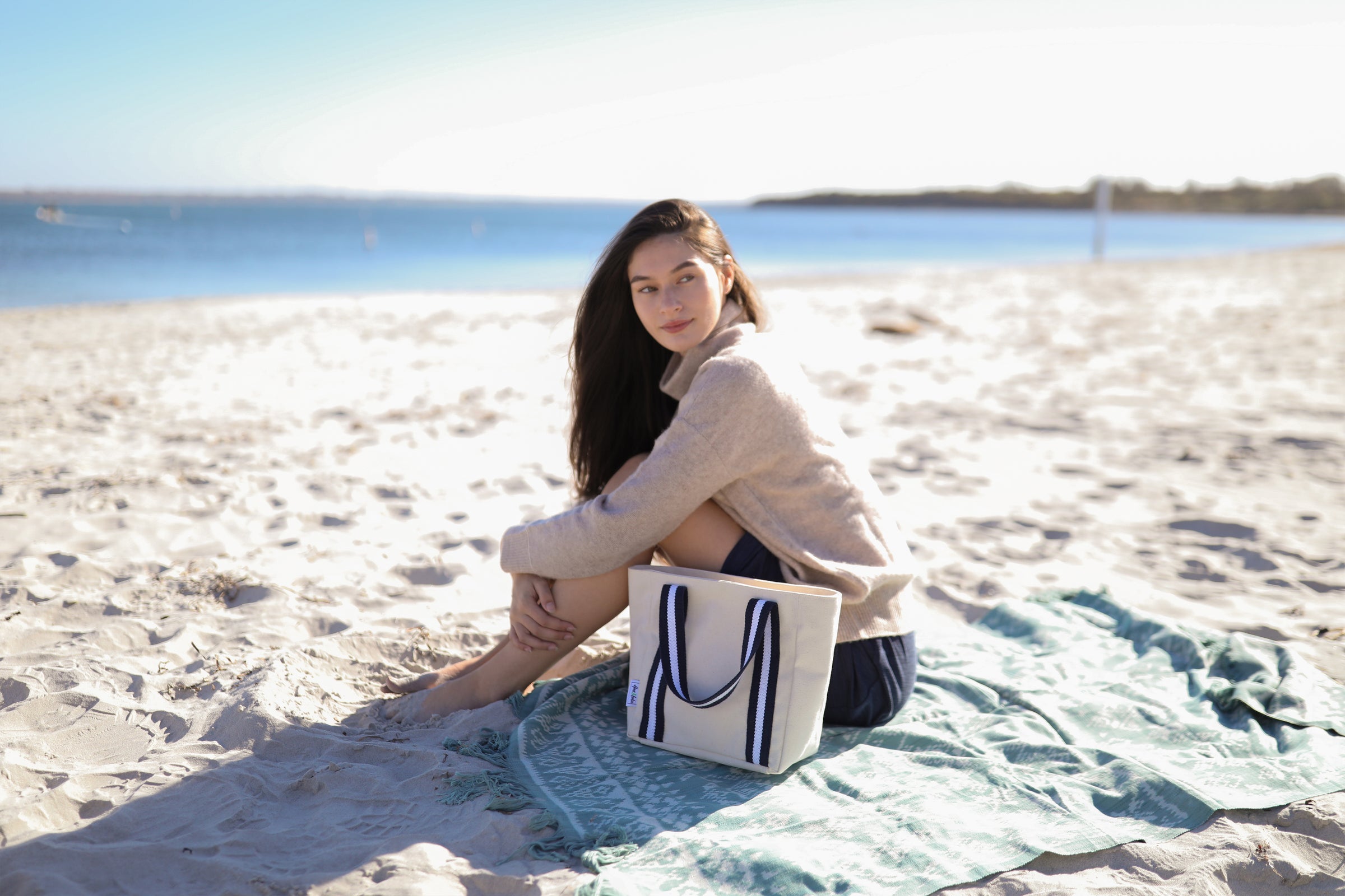 A woman with long brown hair blowing in the wind sits on a beach towel on the beach. There is sand around here and the ocean behind her. On the beach towel in front on her is a small canvas beach tote with navy and white handles.