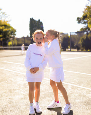 Two young girls stand together on a tennis court. One girl whipsers into the ear of the other girl. Both are wearings white sweatshirts which has Tennis star in handwritten purple font on the front.