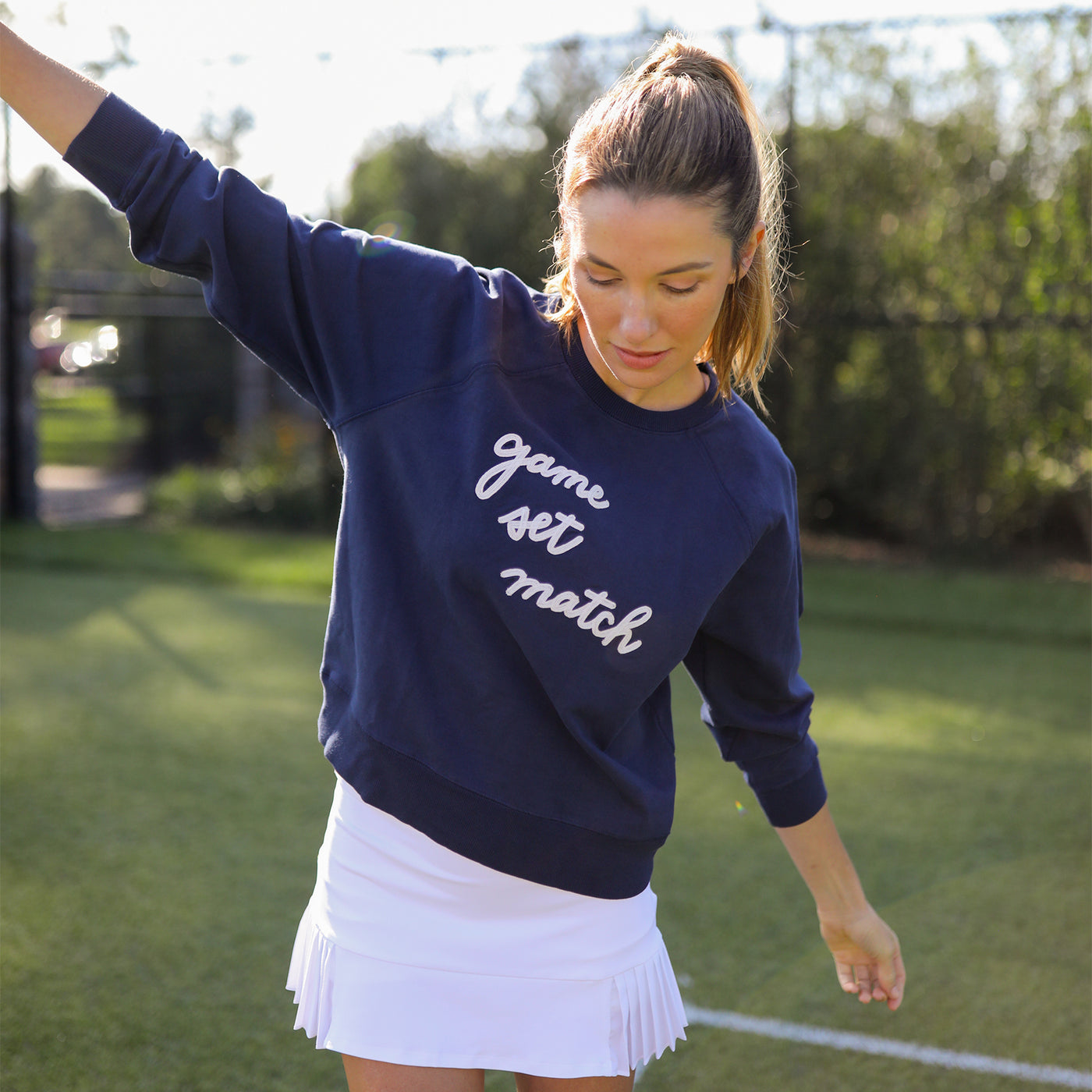 woman stands on tennis court wearing Navy womens sweatshirt lays flats with the text game set match in white cursive font across the front of sweatshirt