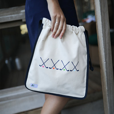 woman holding tan canvas drawstring shoe bag with navy crossed golf clubs on front.