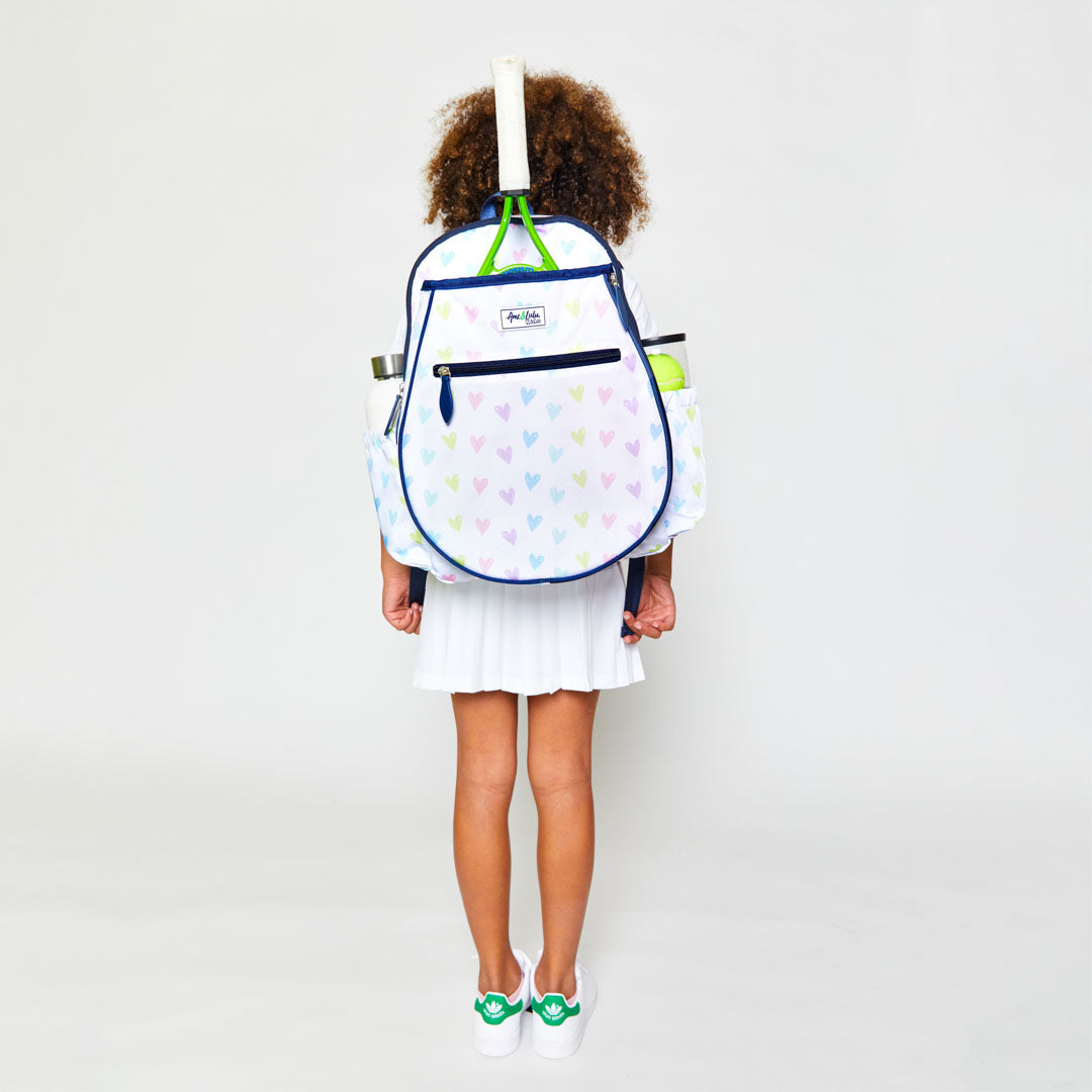Little girl wearing white kids tennis backpack with repeating pattern of rainbow hand drawn hearts.