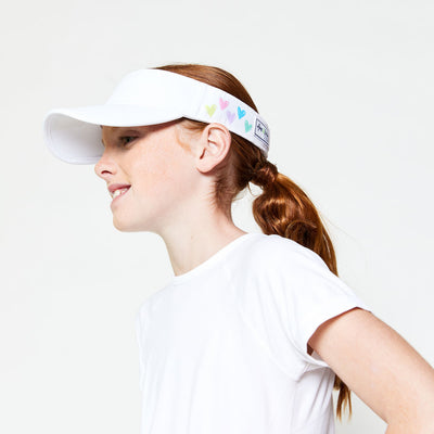 Little girl wearing white kids visor with rainbow hand drawn hearts printed on sides.