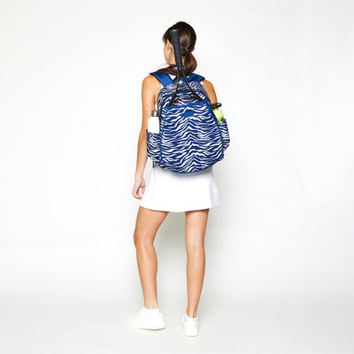 Woman stands on a white background wearing a navy and white tiger pattern tennis backpack.