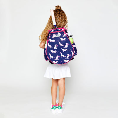 little girl wearing navy kids tennis backpack with hot pink trim and pattern of white puppies holding tennis racquets and tennis balls