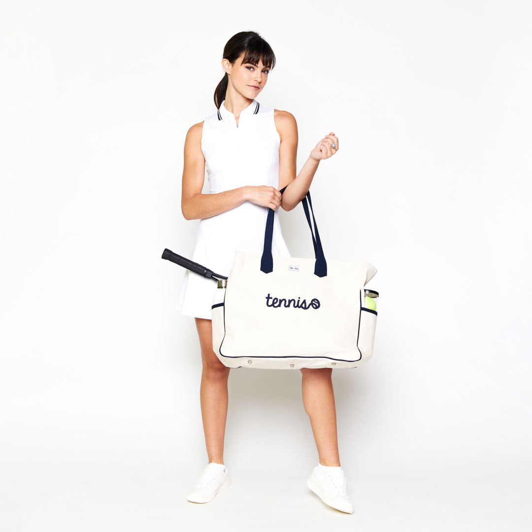 Woman stands on white background holding canvas tote bag. Bag has embroidery on the front that says tennis in a cursive font.