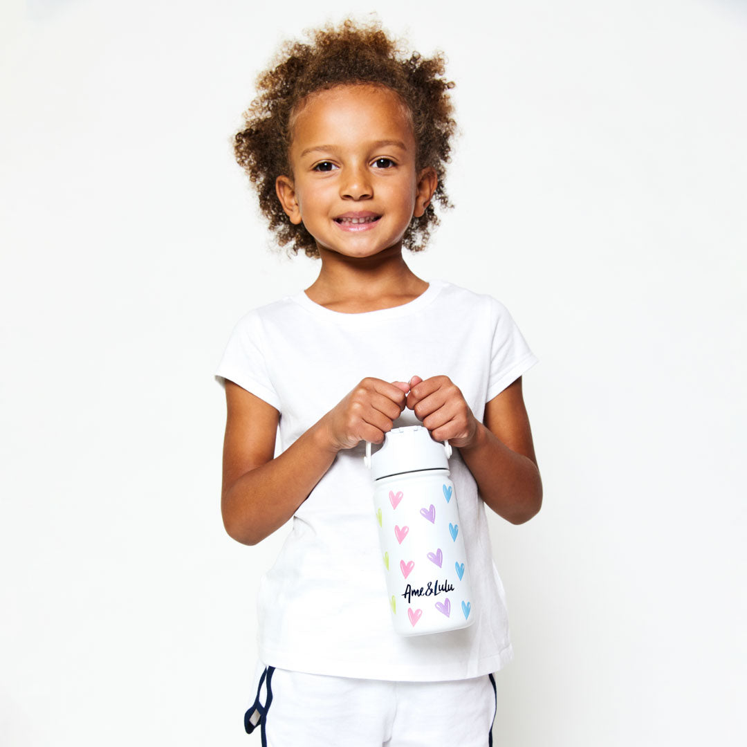 Little girl holding White kids water bottle with rainbow hearts pattern.