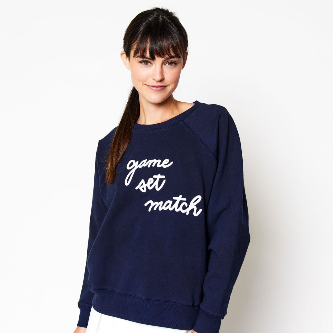 woman stands on white background wearing Navy womens sweatshirt lays flats with the text game set match in white cursive font across the front of sweatshrit