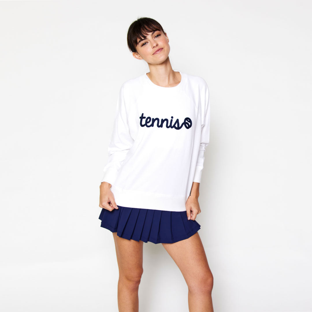 Woman stands on a white background and wears a white sweatshirt. There is navy text that reads tennis in a cursive font across front of sweatshirt
