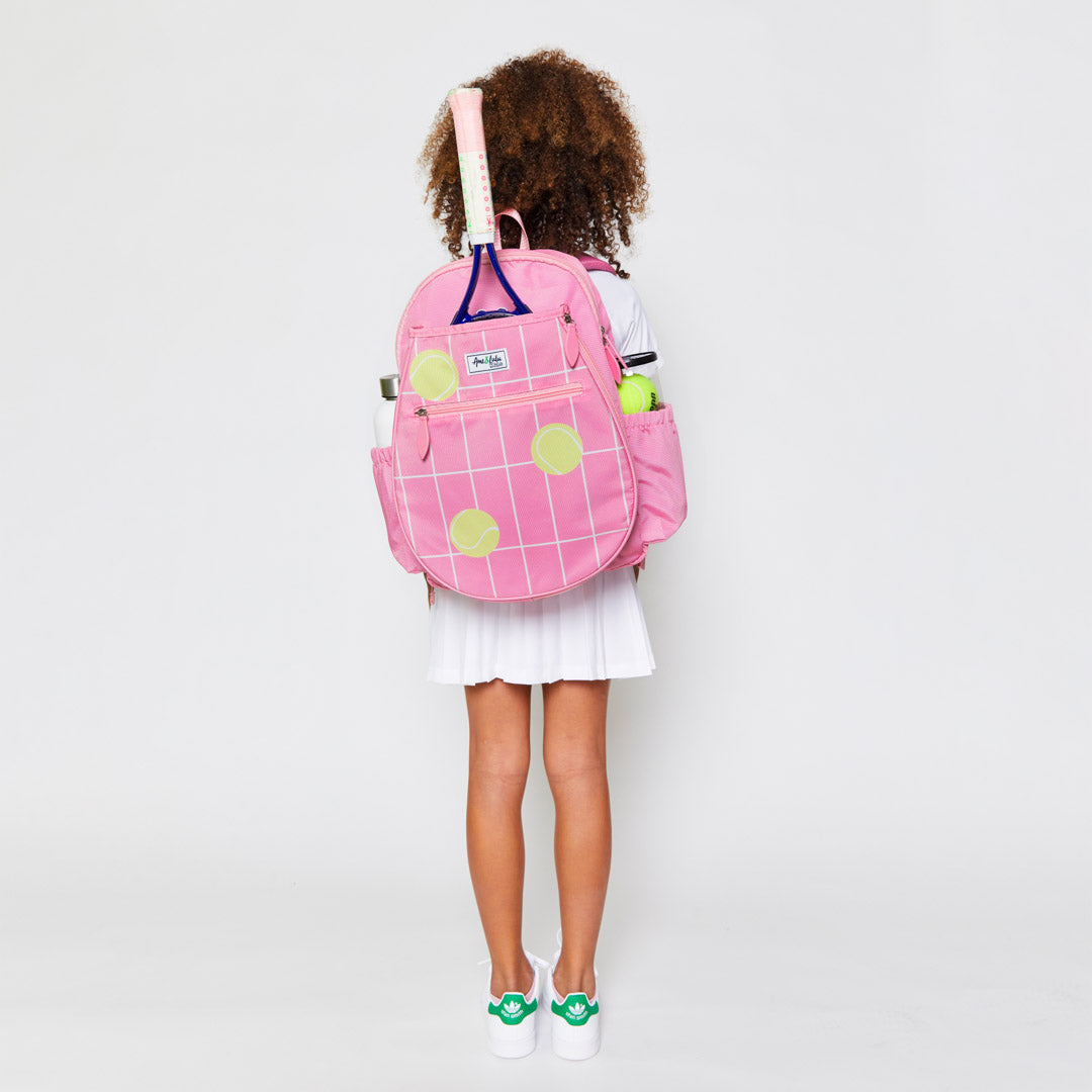 Little girl wearing a coral kids tennis backpack with repeating pattern of cream grid lines and yellow tennis balls.